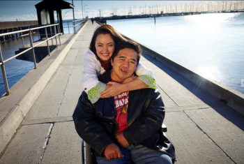 Male NDIS participant on a pier in a wheelchair. His wife is standing behind him with her arms wrapped around him. Both are smiling at the camera.