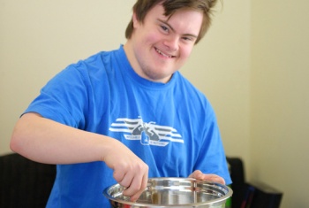 Self-employed 20-year-old in a blue t-shirt, smiling at the camera, using a spoon to mix a cake in a stainless steel mixing bowl.