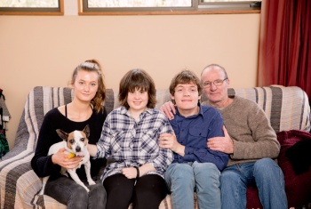 A young female and a young male, who are both NDIS participants and brother and sister, sit in between their dad and their younger sister snuggled on a three-seater couch. The youngest sister has a Jack Russell Terrier, the family pet, sitting on her lap.