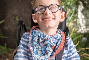 A young boy with glasses sits in a wheelchair smiling