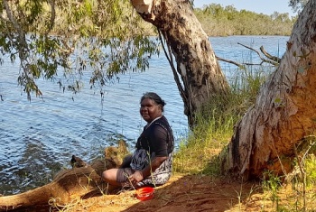 Gaylene sitting on the bank of a river under a tree looking at the camera.