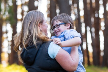 Little boy wearing glasses smiling at his mother as she holds him to her, against a backdrop of blurred out trees.