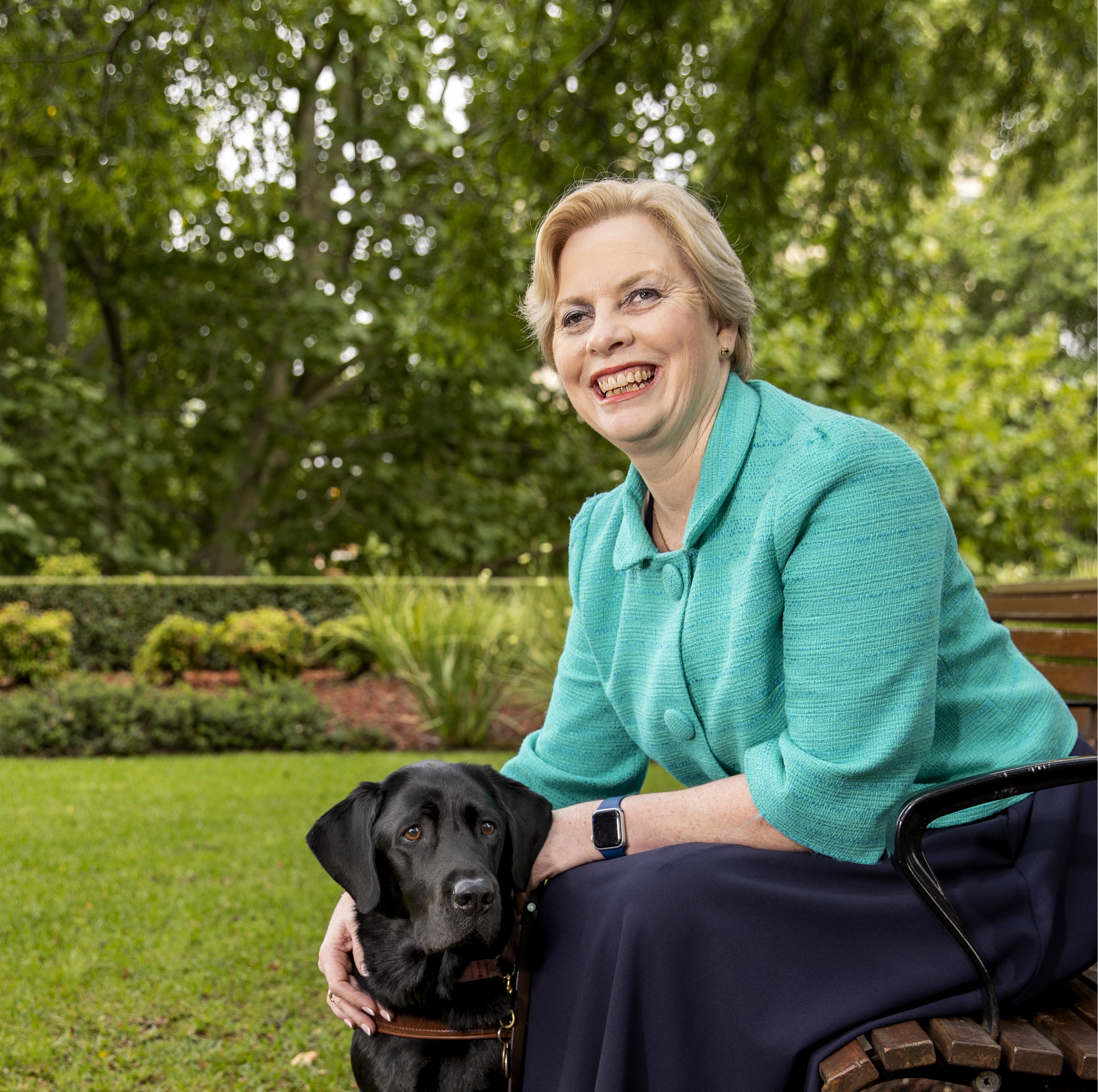 A woman is sitting on a park bench smiling with her guide dog