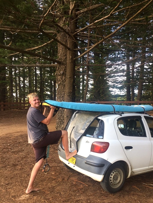 Lleyton unloads stand up paddle board from the roof of his car, he is barefoot, under towering Norfolk Island pine trees