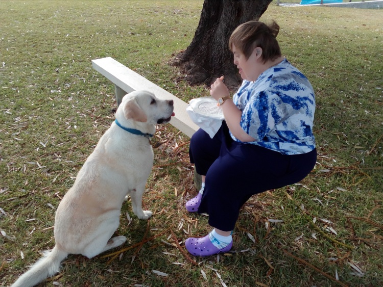 Cathy Sits on a park bench, a labrador sits at attention in front of her, hoping for a treat