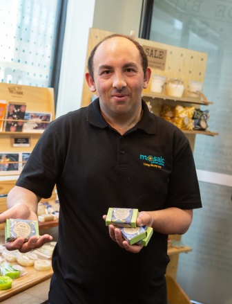 A man with brown hair and a black collared polo top is holding up 4 bars of green soap for the camera. Behind him is a shelf with cards, candles and wheat packs.