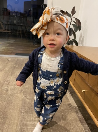 A toddler standing up wearing blue overalls with white flowers and a large headband with yellow flowers. She is looking up at the camera