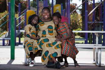 A woman in a colourful African print dress is sitting on a playground smiling with two young girls wearing matching dresses. 