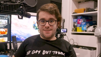 A man with glasses is sitting in a room with recording equipment visible in the background