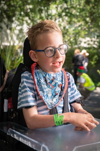 A young boy with glasses sits in a wheelchair outside