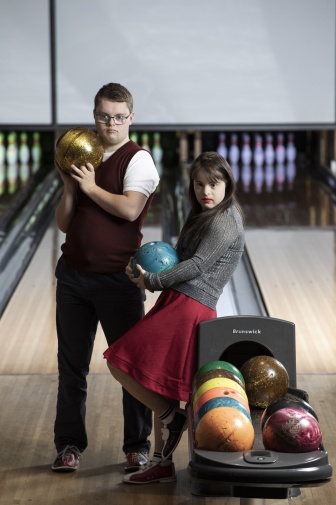 Charlie and Jianna holding bowling balls while standing on a bowling alley, pins are visible in the background.