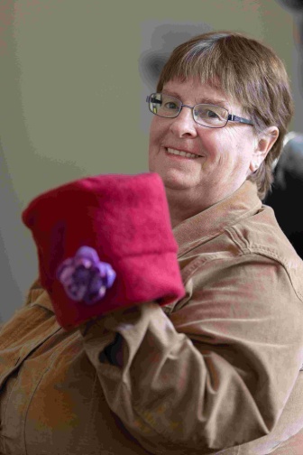 Sharon, sitting, is holding up a red hat with a purple flower she made.