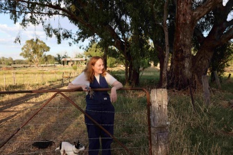 Marli in overalls, her arms over the fence as she smiles, against a backdrop of  trees and blue sky.
