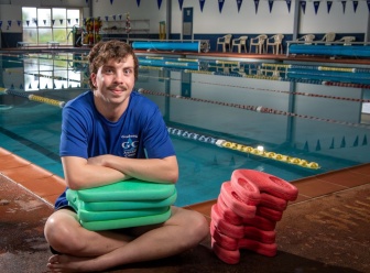 Jonathan sitting cross legged next to a swimming pool with a stack of kickboards under his crossed arms.