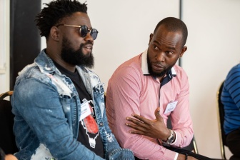 Sengo wearing a denim jacket and sunglasses sitting next to a man in a red shirt who is talking to him. They're both wearing event style name badges.