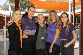 5 women stand outside with their arms around each other looking at the camera