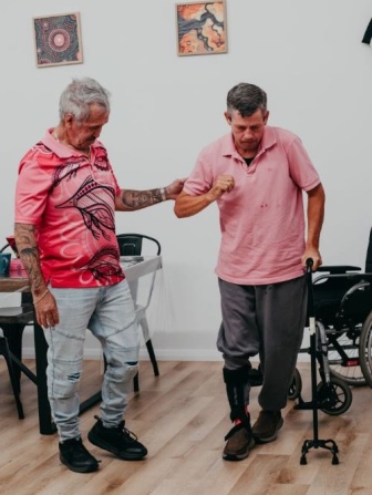 Man in pink shirt and black trousers takes a step forward, assisted by a support worker who supports his arm. He also has a walking stick and leg brace.