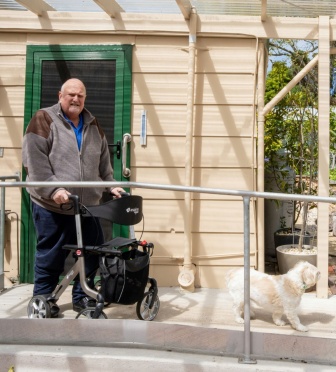 Man stands using a walker at his back door, about to follow his dog down a ramp.