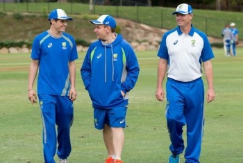 Three men wearing blue cricket uniforms walk across a cricket oval toward the camera.