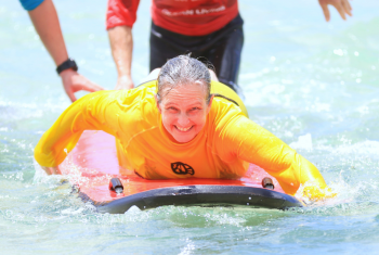 Older female NDIS participant on a surfboard at the beach with support workers coming up behind her.