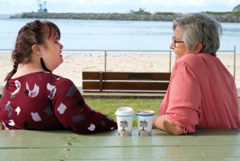 Two women, one with disability, sitting on a park bench at the beach enjoying the view, a chat and a coffee.