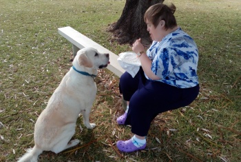 Cathy Sits on a park bench, a labrador sits at attention in front of her, hoping for a treat