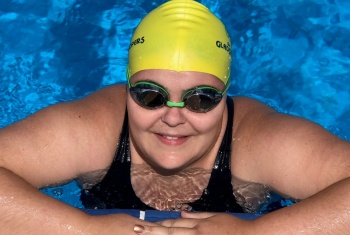 Ruby, in a yellow swimming cap and goggles, is in the pool, her arms folded on the edge, smiling at the camera