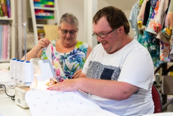 Steven is wearing a white t-shirt and sitting at a sewing machine. A woman in a brightly coloured t-shirt is sitting next to him