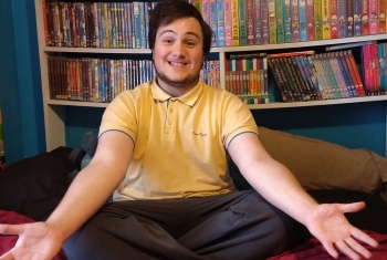 Zac sits cross-legged in front of shelves sagging under the weight of DVDs and videos. He has a huge smile and open arms.