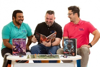 Three men are sitting at a table with books about the game Dungeons and Dragons. The man in the middle is holding his book open and smiling widely, the men on each side are facing inward to look at the middle man.