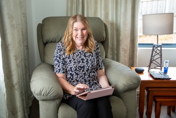 A woman with shoulder length blonde hair is sitting in a large armchair holding an ipad.