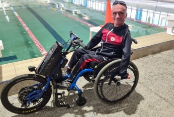 A man in a three wheeled wheelchair is smiling at the camera with an indoor pool in the background.