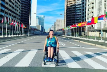A woman wearing an Australian Olympics top is in a wheelchair in the middle of an empty road. On either side of the road are rows of flags from around the world.