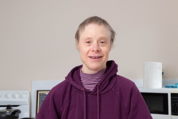 An older woman with down syndrome stands smiling in a kitchen holding a cup of tea.