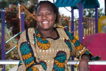 A woman wearing a colourful African print dress is sitting on a playground smiling.