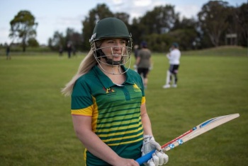 A woman in a sports uniform is standing on a field wearing a batting helmet and holding a cricket bat.