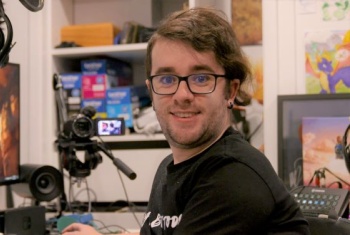 A man with glasses is sitting at a computer with recording equipment visible in the background.