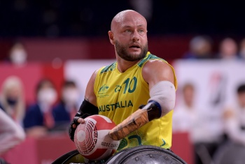 A man in a wheelchair wearing an Australian Olympic singlet holds a ball, about to throw it. In the background is a blurred crowd watching.