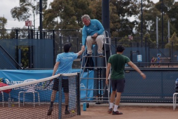 Male tennis umpire sitting atop highchair shakes hands with players
