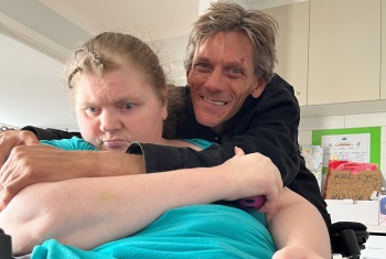 Woman in wheelchair being hugged by her smiling husband. A bag in the background is labelled 'Kelly's pampering equipment - please use"