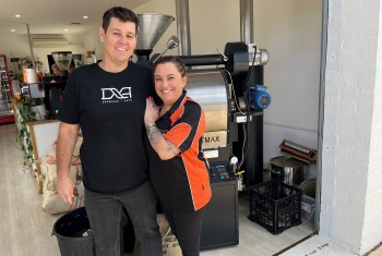 Tamina and her husband smiling as they pose in the cafe they manage. A coffee machine and staff can be seen in the background.