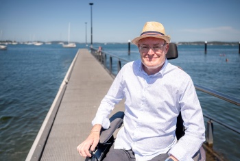 Man sitting on motorised chair in shirt and hat, on a jetty on a sunny day