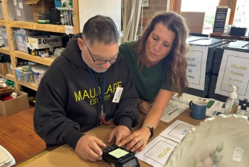 Man in black hoodie looking at trader EFTPOS machine, while woman shows him how to use it.