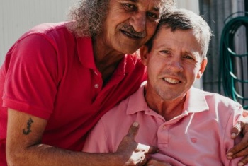 Man in pink shirt sitting, being hugged side on by a man in dark pink shirt.