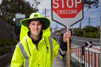 Man in high-visibility coat and hat holds a STOP sign. He is standing beside a school crossing.