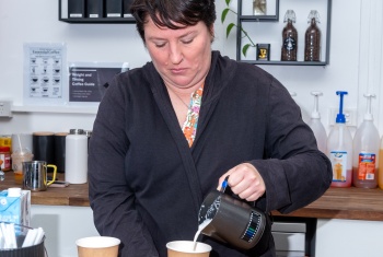 Elise stands behind the counter at a cafe pouring a coffee