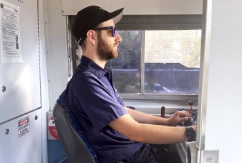 Josh sits in the drivers seat of a train wearing a uniform. He is looking forwarded, focused.